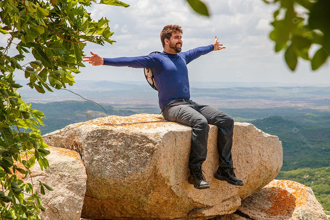 homem de braços abertos, sentado na rocha em uma montanha. Conceito de aventura e vitória
