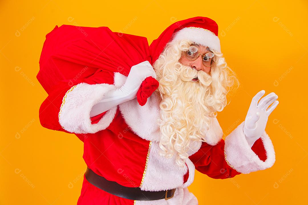 Papai Noel em fundo amarelo, segurando o saco com presentes nas costas. Papai Noel fazendo uma surpresa na noite de Natal