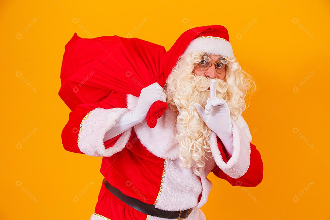 Papai Noel em fundo amarelo, segurando o saco com presentes nas costas. Papai Noel fazendo uma surpresa na noite de Natal