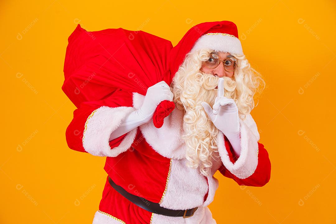 Papai Noel em fundo amarelo, segurando o saco com presentes nas costas. Papai Noel fazendo uma surpresa na noite de Natal