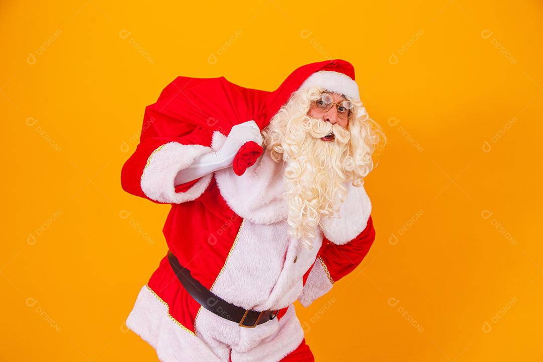 Papai Noel em fundo amarelo, segurando o saco com presentes nas costas. Papai Noel fazendo uma surpresa na noite de Natal