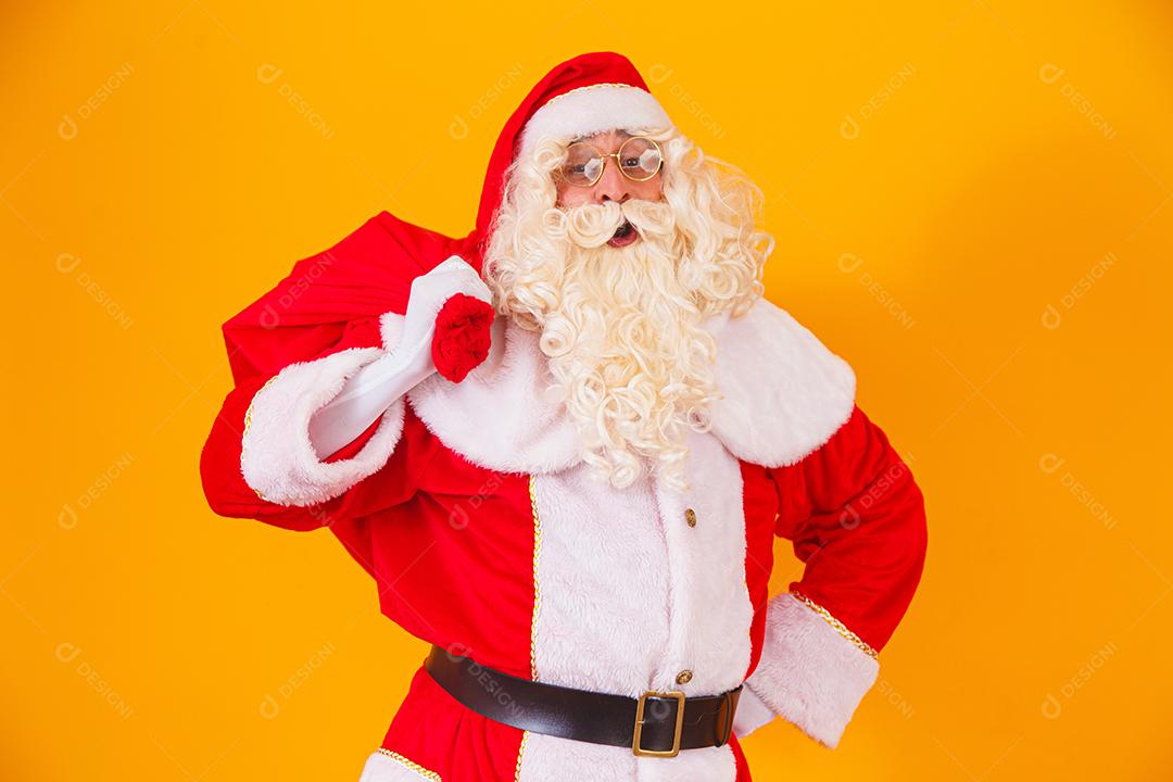 Papai Noel em fundo amarelo, segurando o saco com presentes nas costas. Papai Noel fazendo uma surpresa na noite de Natal