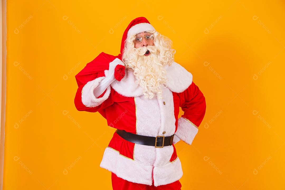 Papai Noel em fundo amarelo, segurando o saco com presentes nas costas. Papai Noel fazendo uma surpresa na noite de Natal