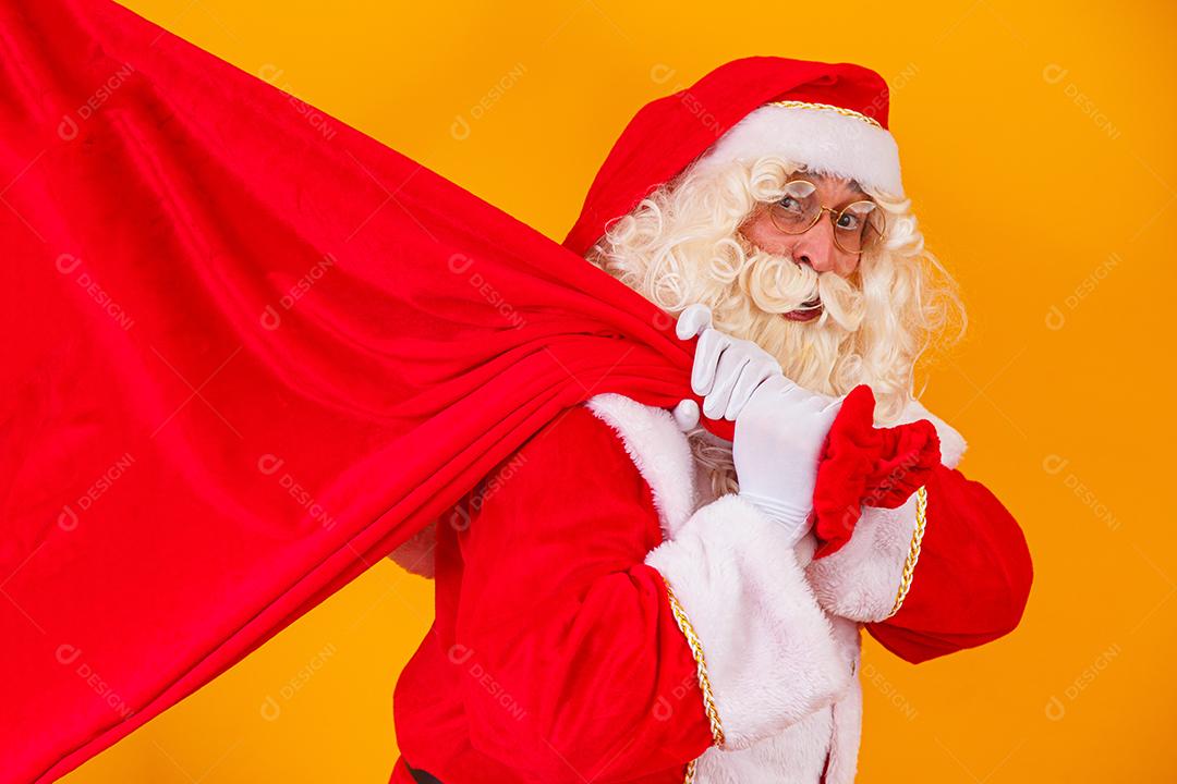 Papai Noel em fundo amarelo, segurando o saco com presentes nas costas. Papai Noel fazendo uma surpresa na noite de Natal