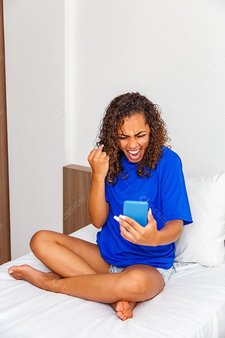 Jovem mulher no quarto usando seu celular comemorando.