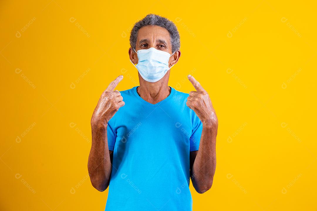 Homem idoso usando mascara branca apontando para si em fundo amarelo