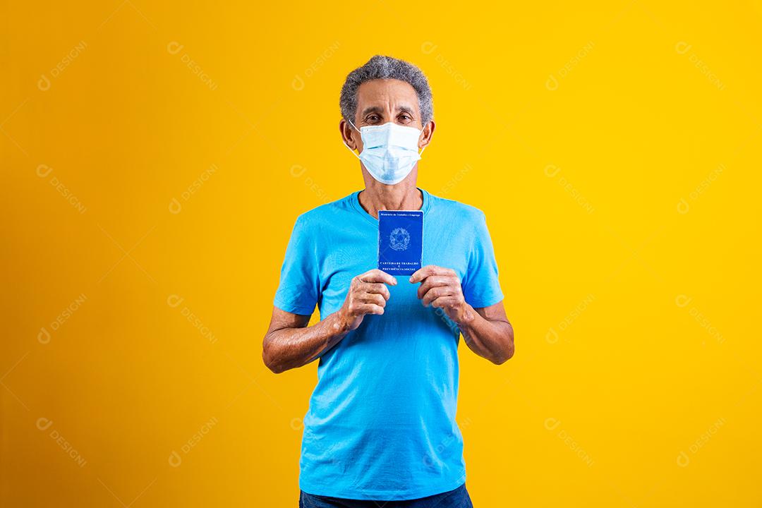 Homem idoso usando mascara branca e segurando carteira de trabalho em fundo amarelo