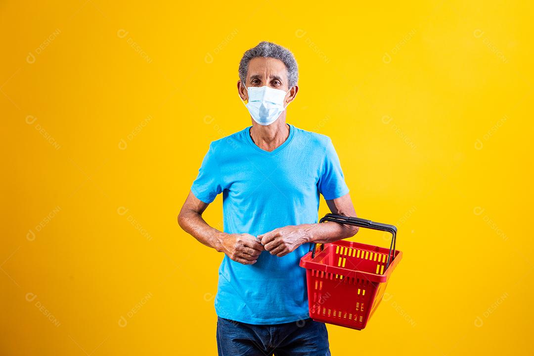 Homem idoso usando mascara branca e segurando cesta de mercado em fundo amarelo