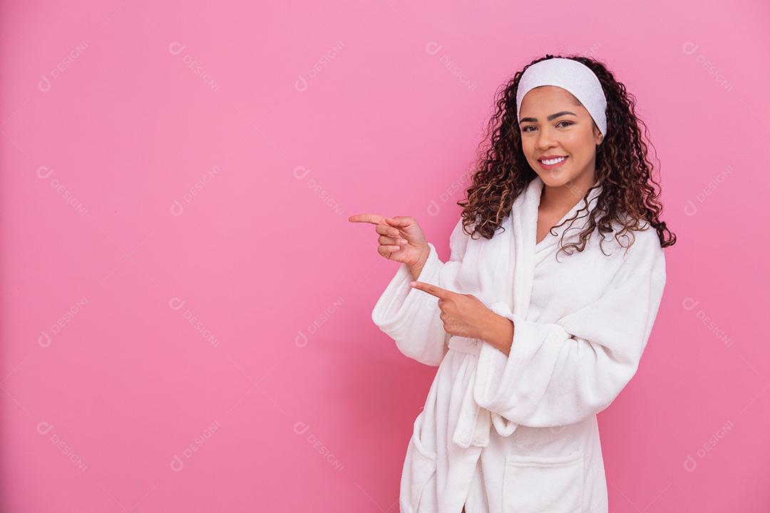 Linda mulher afro, vestida com um roupão de banho, apontando para o lado no fundo rosa. mulher afro vestido com poses de manto branco fofo