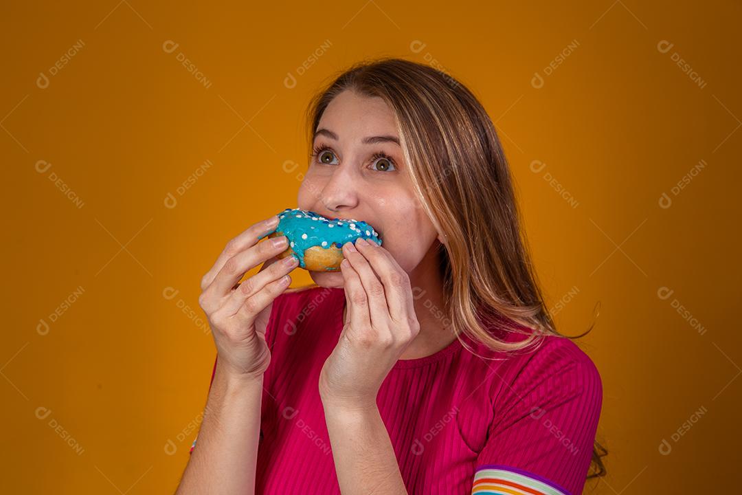 Retrato de uma jovem loira comendo uma deliciosa rosquinha colorida.
