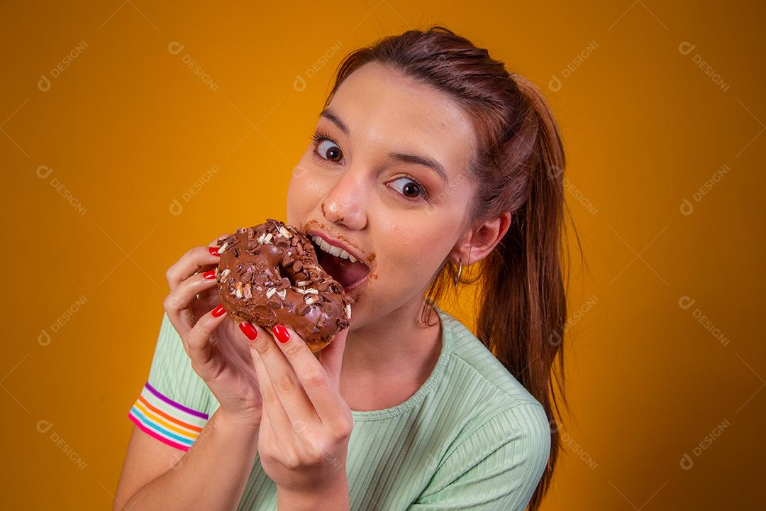 Jovem ruiva comendo deliciosos donuts de chocolate com a boca manchada com cobertura.