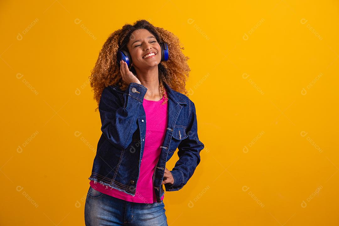 Mulher jovem e bonita com cabelo afro ouvindo música com seu fone de ouvido em fundo amarelo