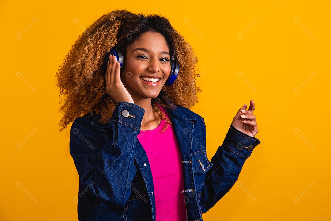 Mulher jovem e bonita com cabelo afro ouvindo música com seu fone de ouvido em fundo amarelo