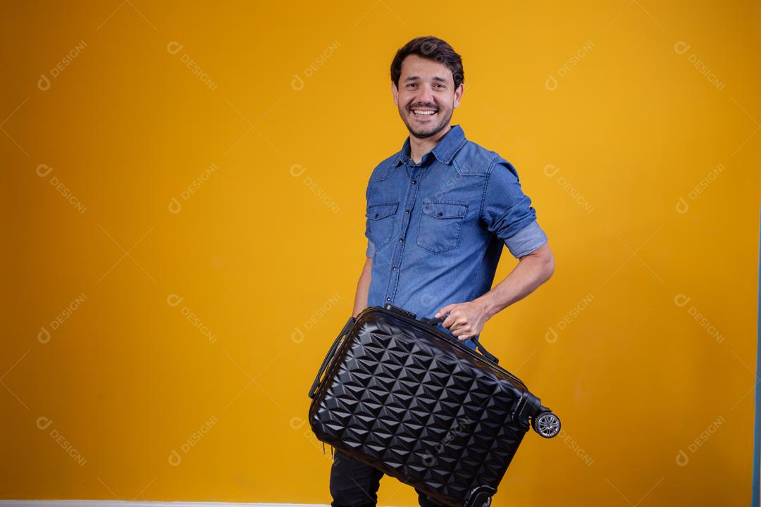 Homem com camisa jeans em fundo amarelo segurando mala preta