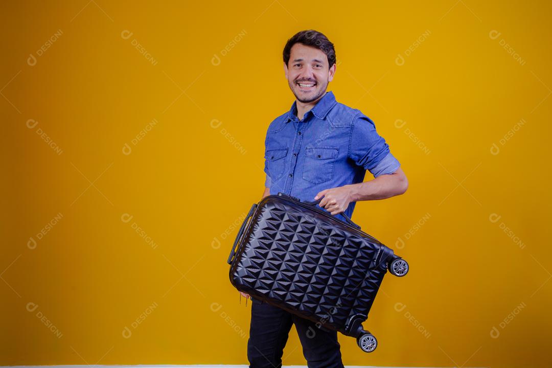 Homem com camisa jeans em fundo amarelo segurando mala preta