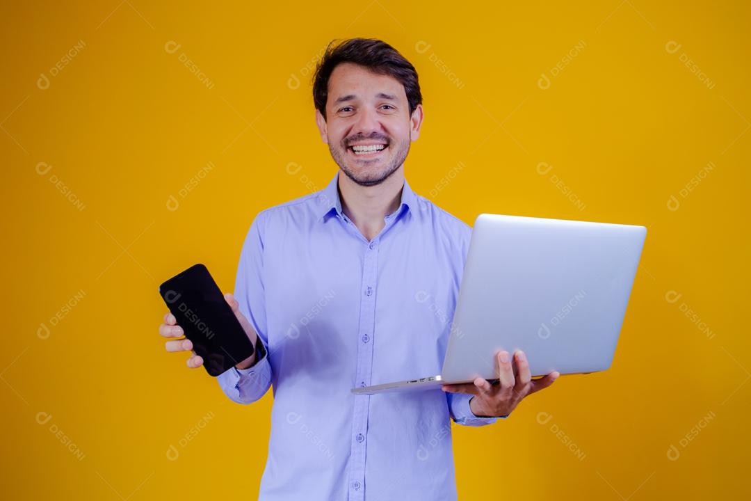 Homem com camisa social manga longa em fundo amarelo Segurando celular e Notebook e Computador