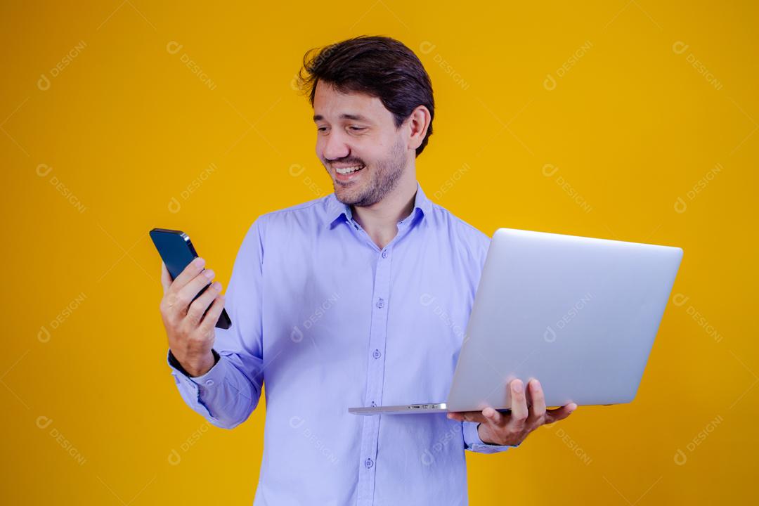 Homem com camisa social manga longa em fundo amarelo Segurando celular e Notebook e Computador
