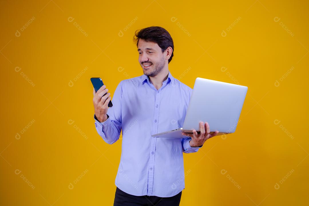 Homem com camisa social manga longa em fundo amarelo Segurando celular e Notebook e Computador