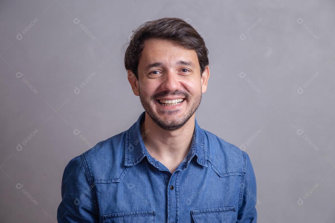Homem  com roupa social camisa jeans e expressão de felicidade em fundo cinza