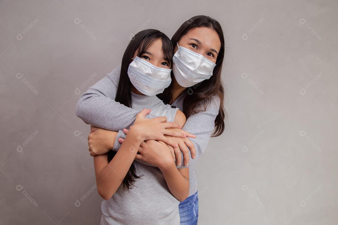 Mãe e filha abraçadas com mascaras em fundo bege