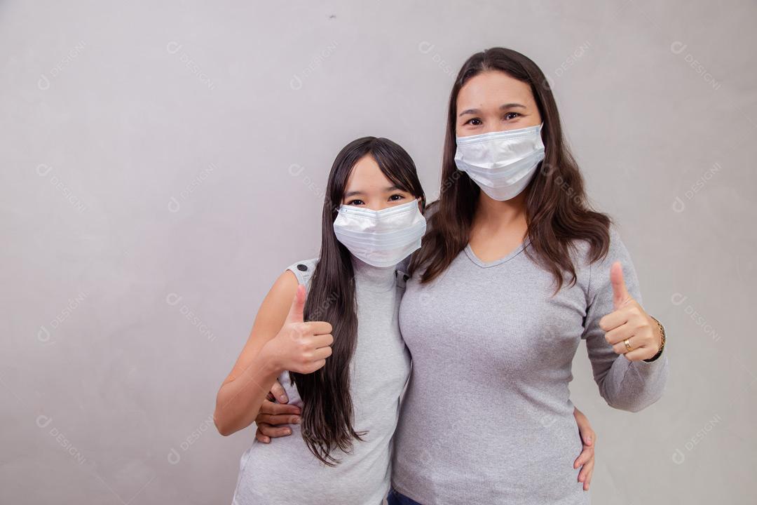 Mãe e filha abraçadas com mascaras em fundo bege