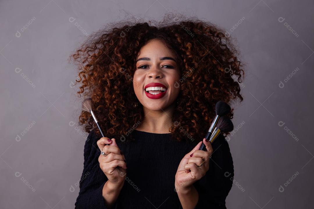 Mulher com cabelo cacheado maquiadora com pinceis de maquiagem na mão em fundo cinza