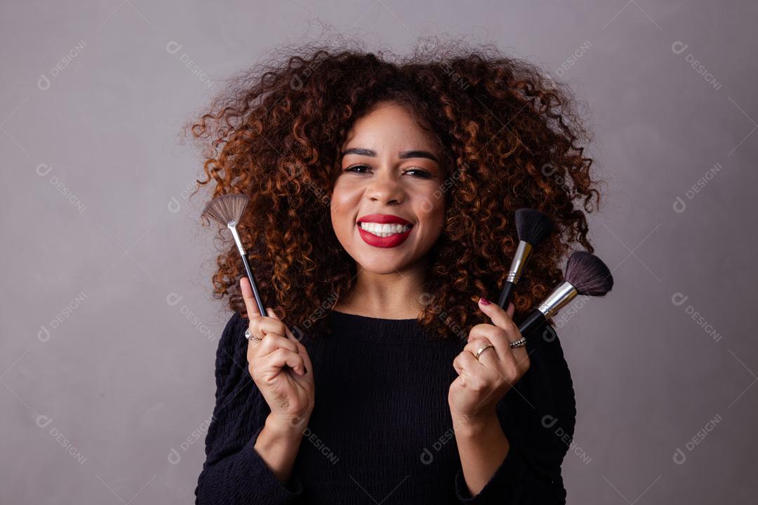 Mulher com cabelo cacheado maquiadora com pinceis de maquiagem na mão em fundo cinza