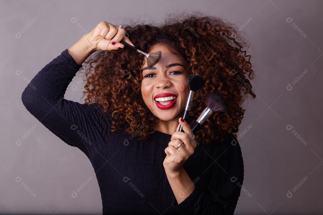 Mulher com cabelo cacheado maquiadora com pinceis de maquiagem na mão em fundo cinza