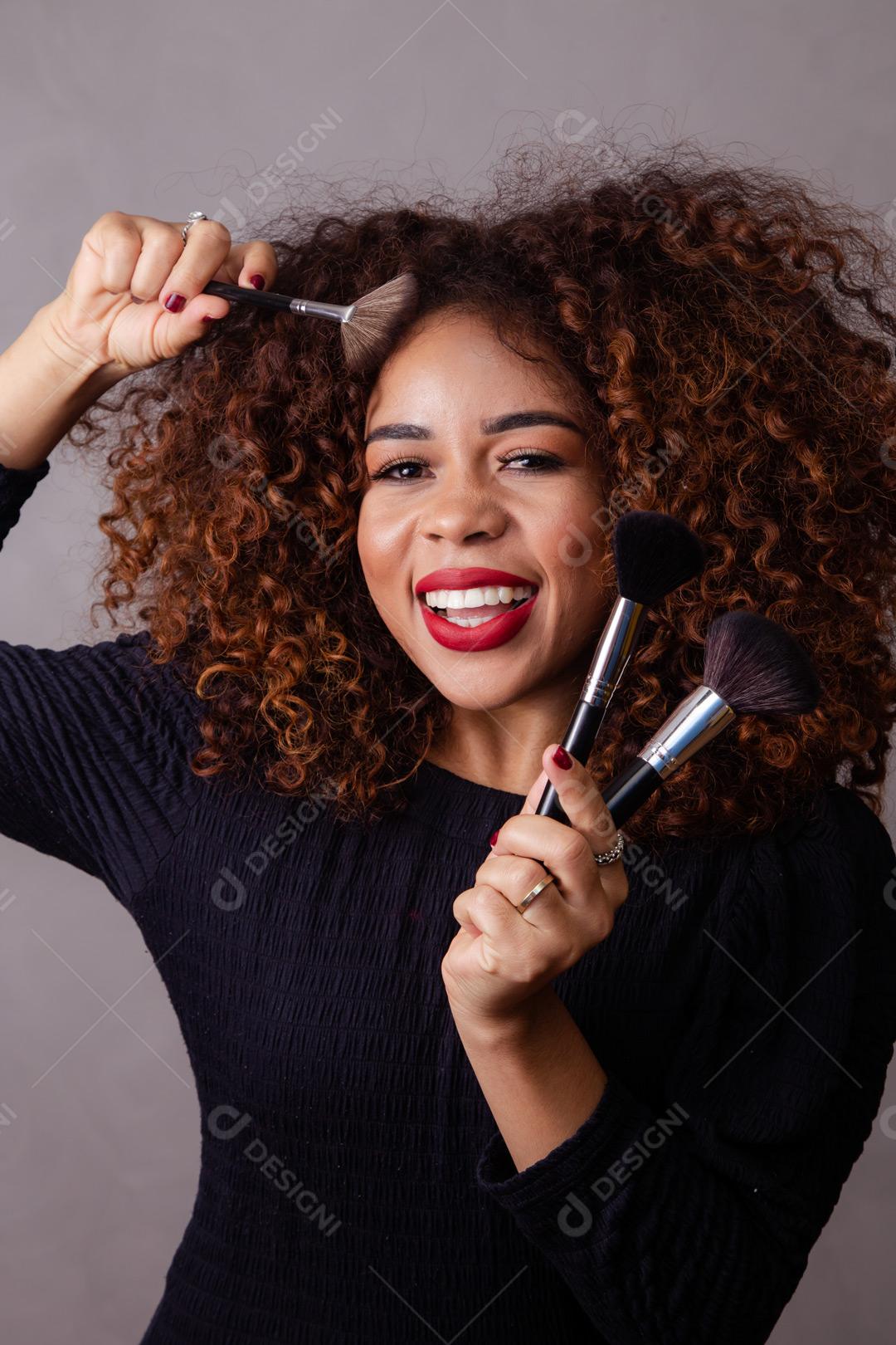 Mulher com cabelo cacheado maquiadora com pinceis de maquiagem na mão em fundo cinza