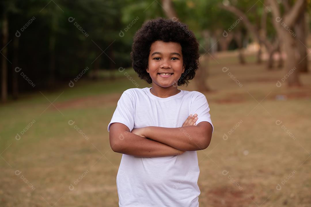 menino afro com os braços cruzados com o parque ao fundo.