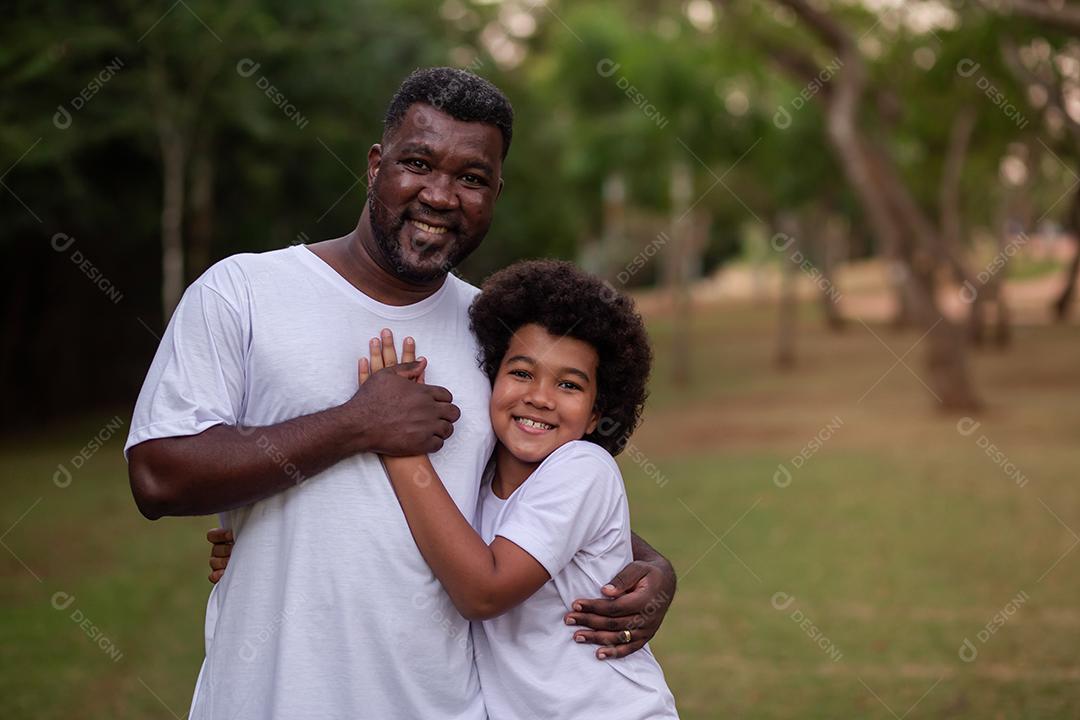 Pai e filho negros no parque abraçados sorrindo olhando para a câmera