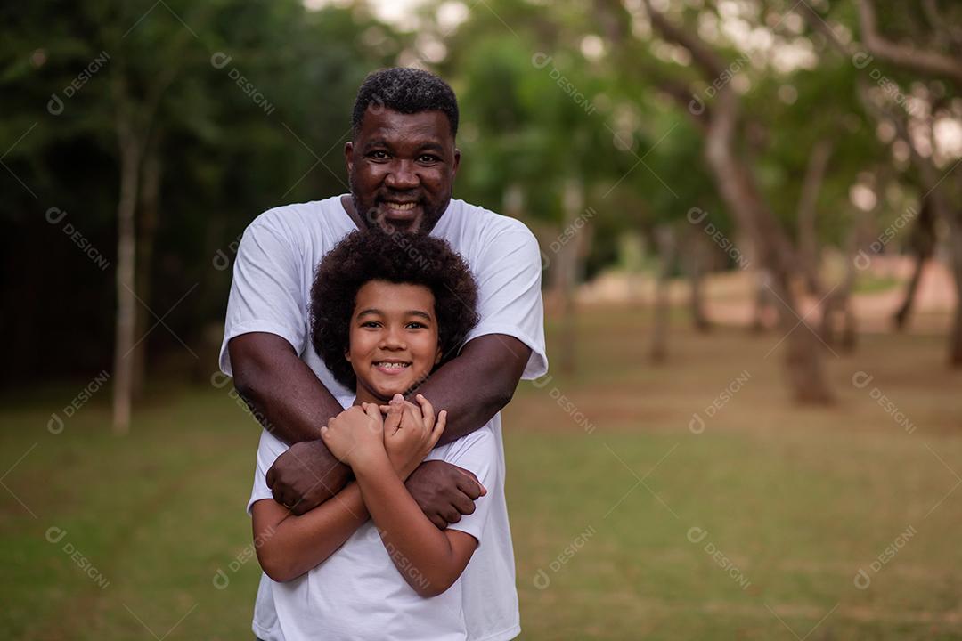 Pai e filho negros no parque abraçados sorrindo olhando para a câmera