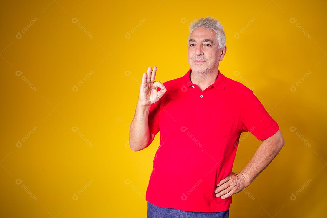 Retrato de homem idoso alegre mostrando o gesto okey isolado em fundo amarelo.