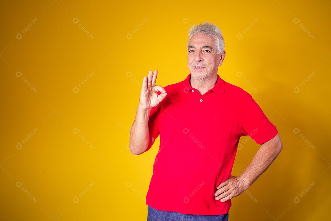 Retrato de homem idoso alegre mostrando o gesto okey isolado em fundo amarelo.