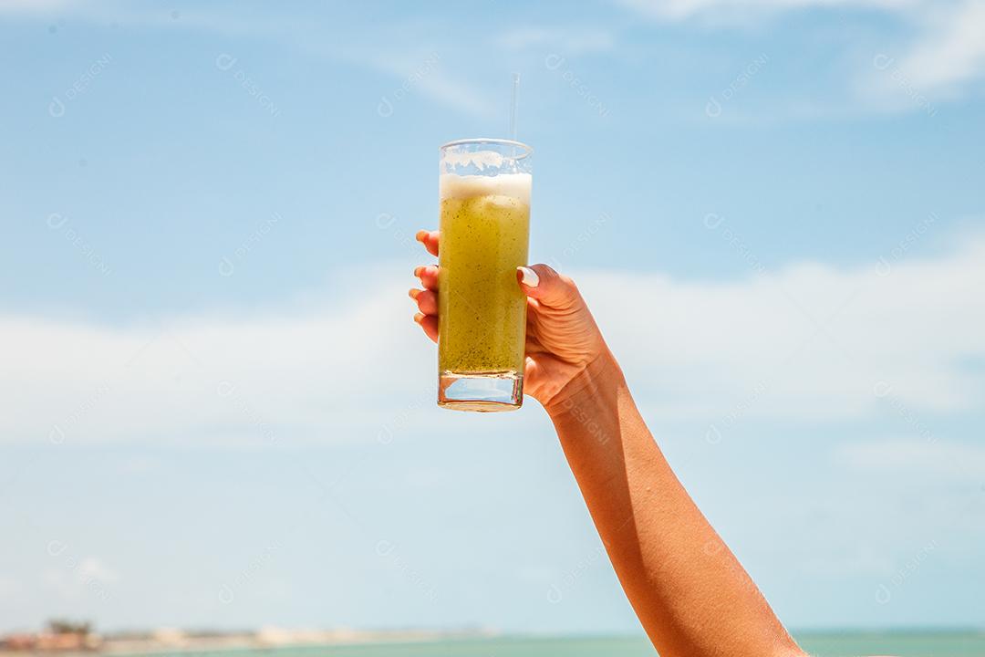 mãos de mulher segurando um suco de abacaxi com a praia ao fundo.