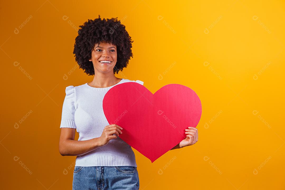 Retrato fotográfico de uma mulher afro sorridente segurando um grande cartão de coração vermelho