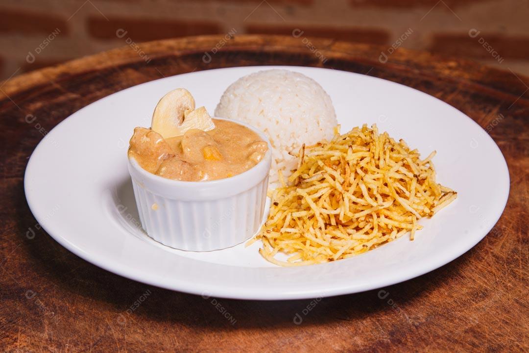 Prato de comida com estrogonofe de frango e arroz e batatas palhas sobre mesa de madeira