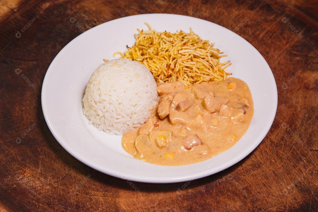 Prato de comida com estrogonofe de frango e arroz e batatas palhas sobre mesa de madeira