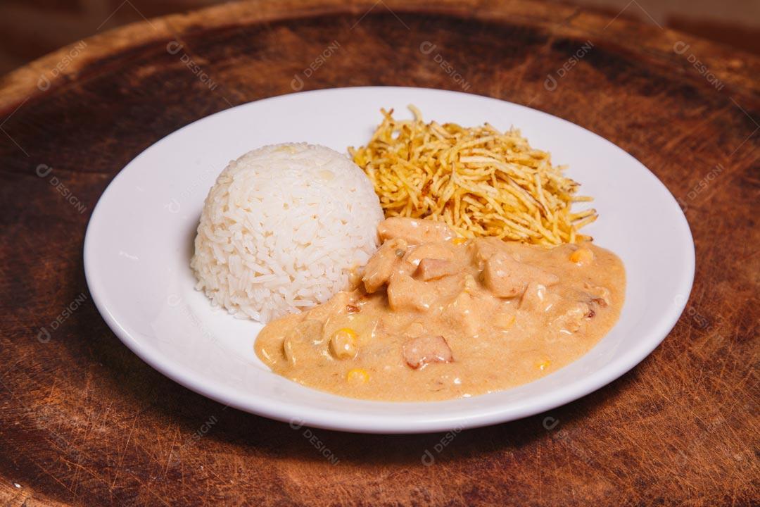 Prato de comida com estrogonofe de frango e arroz e batatas palhas sobre mesa de madeira