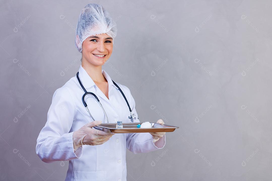 Mulher médica com jaleco e estetoscópio segurando bandeja com remédios em fundo cinza