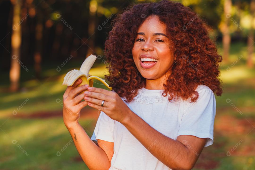 Mulher afro comendo banana no parque. Conceito de vida saudável