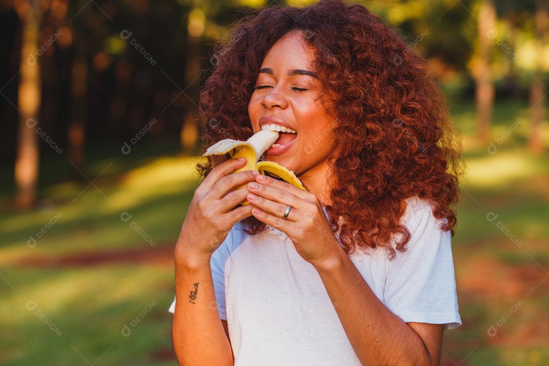 Mulher afro comendo banana no parque. Conceito de vida saudável