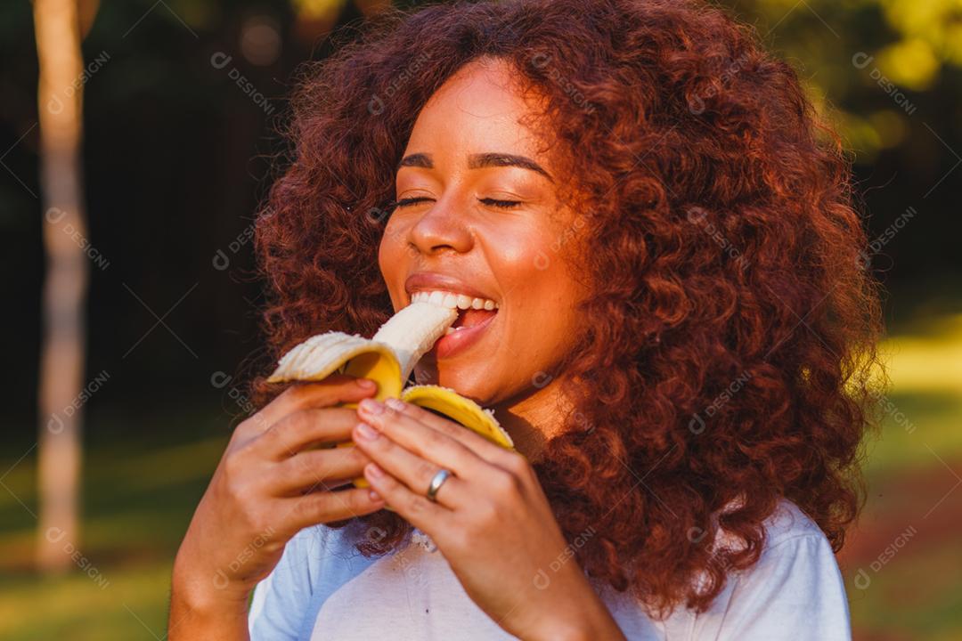 Mulher afro comendo banana no parque. Conceito de vida saudável