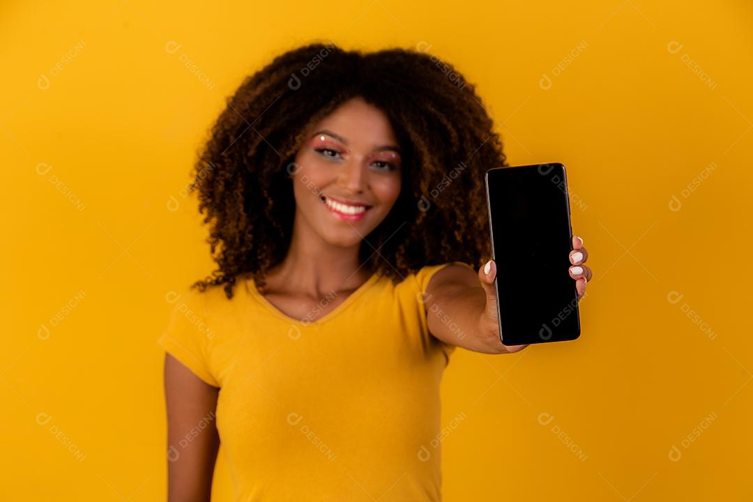 mulher afro com cabelo encaracolado, apontando para o celular em fundo amarelo