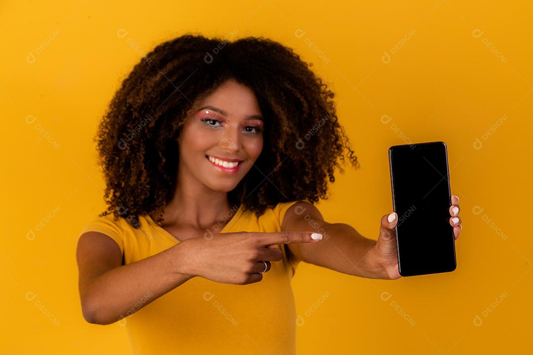 mulher afro com cabelo encaracolado, apontando para o celular em fundo amarelo