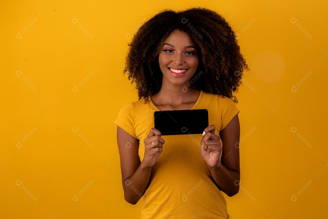 mulher afro com cabelo encaracolado, apontando para o celular em fundo amarelo