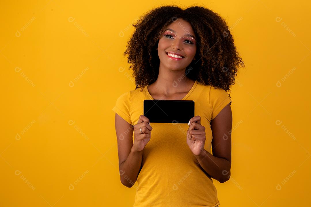 mulher afro com cabelo encaracolado, apontando para o celular em fundo amarelo