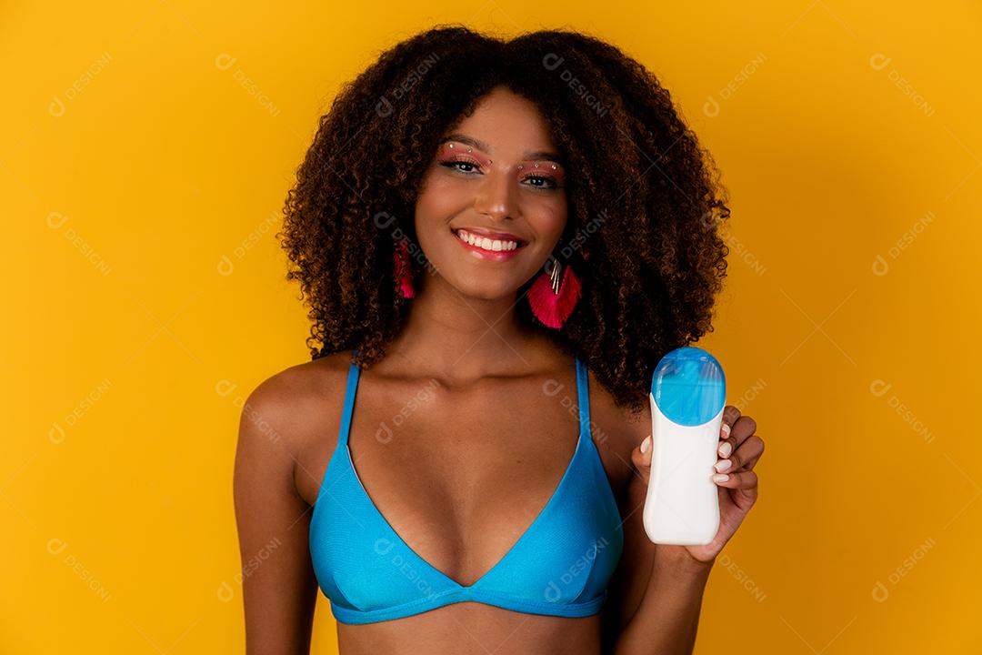 Mulher afro segurando uma garrafa de protetor solar em branco. Apresentando um produto. Conceito de verão, praia e piscina.