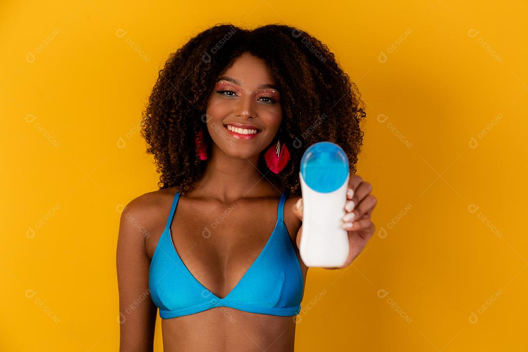 Mulher afro segurando uma garrafa de protetor solar em branco. Apresentando um produto. Conceito de verão, praia e piscina.
