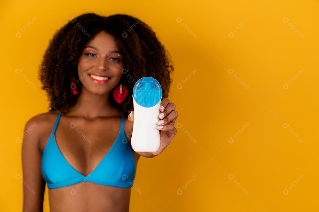 Mulher afro segurando uma garrafa de protetor solar em branco. Apresentando um produto. Conceito de verão, praia e piscina.